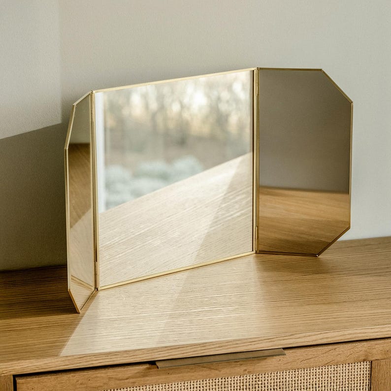 May include: A gold-toned, three-sided vanity mirror with a bevelled edge. The mirror is sitting on a wooden surface.