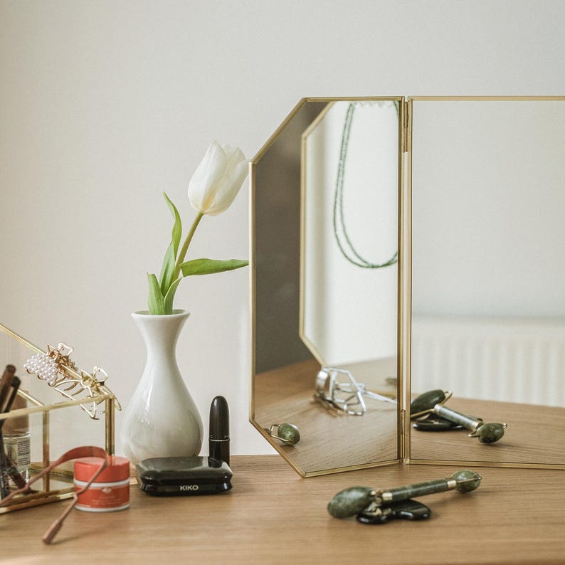 May include: A gold-framed tri-fold mirror with a white tulip in a white vase, a black makeup compact, a black lipstick, and a green jade roller on a wooden surface.
