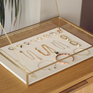 Può includere: Un portagioie rettangolare in vetro con cornice in metallo dorato, contenente un assortimento di anelli, collane, bracciali color oro e argento e un piccolo ciondolo a forma di orsetto su un rivestimento in tessuto color crema.