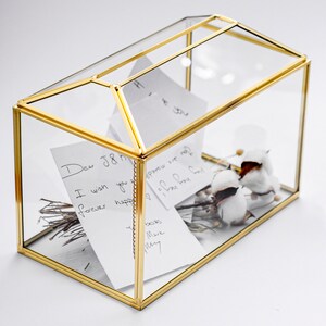 May include: A rectangular glass box with a gold metal frame and a gabled roof lid. Inside the container are several white paper notes with handwritten text, including the phrase Dear J&K, along with small dried twigs and white cotton bolls.