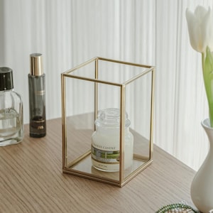 Könnte beinhalten: Ein goldfarbener Kerzenhalter aus Metall mit quadratischer Basis und klaren Glasseiten. Ein weißes Kerzenglas mit dem Text "Yankee Candle Clean Cotton" befindet sich im Halter.