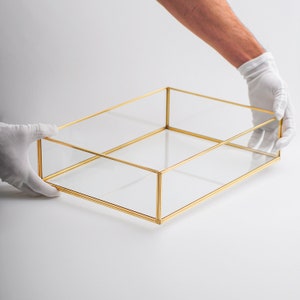 Könnte beinhalten: Eine transparente Glasbox mit goldenem Metallrahmen. Die Box ist leer und wird von zwei behandschuhten Händen gehalten.