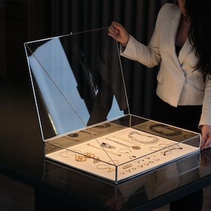 Może przedstawiać: Przezroczysta szklana gablota z otwieraną pokrywą, prezentująca różne naszyjniki, bransoletki i kolczyki. Biżuteria ułożona jest na białej powierzchni, oświetlonej wewnętrznym światłem. Gablota stoi na ciemnej, odbijającej światło powierzchni.