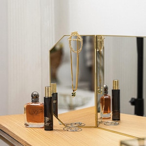May include: A gold-framed, three-panel mirror with a gold necklace hanging in the centre panel. Two bottles of perfume are on the table in front of the mirror.