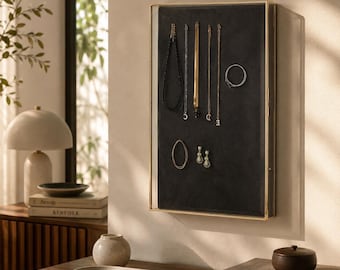May include: A rectangular wall-mounted jewelry display case featuring a thin gold metal frame and a dark charcoal gray velvet interior. The case holds several necklaces, a circular bracelet, and a pair of drop earrings arranged on the surface.