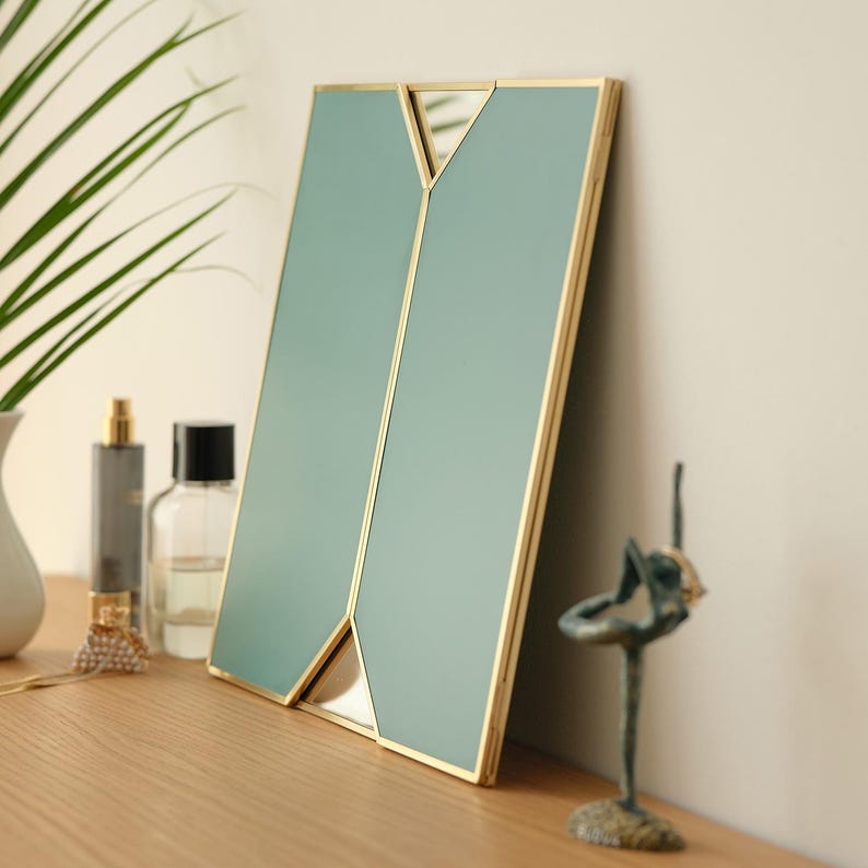May include: A decorative mirror with a gold frame and a light blue glass surface. The mirror is divided into two sections by a gold geometric design.