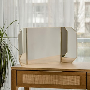 May include: A gold-framed, three-panel vanity mirror with a wooden vanity table with a woven wicker drawer.