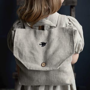 以下が含まれることがあります： 前面に小さな黒い鳥が刺繍された薄いグレーのリネン製のバックパック。バックパックは木製のボタン留めのあるフラップ付きです。
