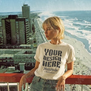 Puede incluir: Camiseta color crema con el texto "YOUR DESIGN HERE" en azul oscuro. La persona lleva la camiseta con las mangas dobladas. Vista de una persona en un balcón con vistas a la playa y la ciudad.