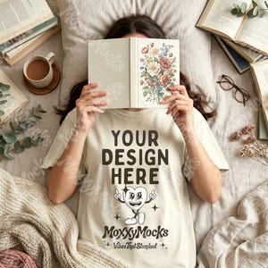 May include: A cream-colored t-shirt with the text "YOUR DESIGN HERE" and a heart-shaped cartoon character. The shirt is part of a mockup, with books, a cup of coffee, and blankets arranged around it. The text "MoxxyMocks" and "Vibes That Standout" are also visible.