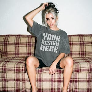 May include: A gray t-shirt with the text "YOUR DESIGN HERE" in white block letters. The person is wearing black shorts and sitting on a plaid couch with red and tan tones. The background is a plain white wall.