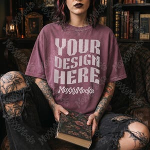 May include: A maroon t-shirt with the text "YOUR DESIGN HERE" in white pixelated font. The shirt is worn by a person seated in a leather chair, holding a book with a floral cover. The person is wearing ripped black jeans.
