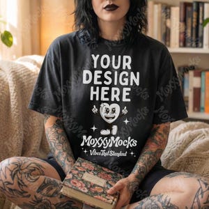 May include: Black t-shirt with the text "YOUR DESIGN HERE" and a heart-shaped graphic. The shirt also features the text "MoxxyMocks" and "Vibes That Standout". The person is holding a book with a floral cover.