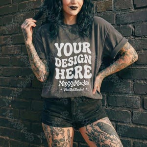 May include: A gray t-shirt with the text "YOUR DESIGN HERE" in white, with "MoxxyMocks" and "Vibes That Standout" below. The model is wearing black shorts and is standing in front of a dark brick wall.