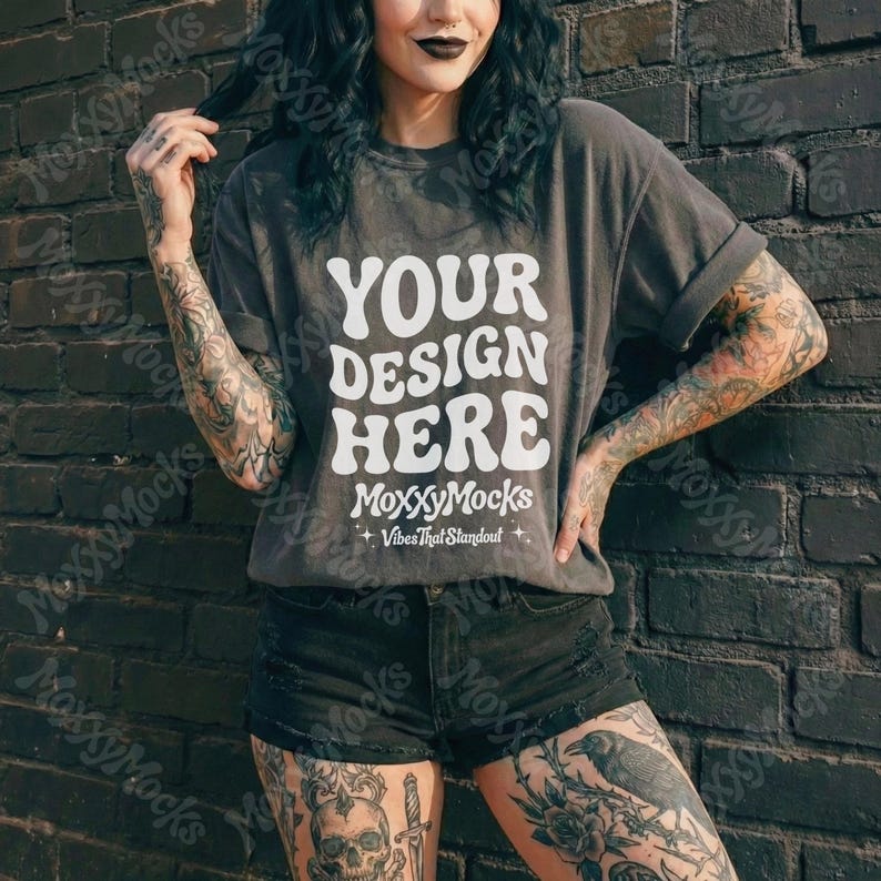 May include: A gray t-shirt with the text "YOUR DESIGN HERE" in white, with "MoxxyMocks" and "Vibes That Standout" below. The model is wearing black shorts and is standing in front of a dark brick wall.