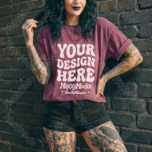May include: A maroon t-shirt with the text "YOUR DESIGN HERE" in a white, retro font. The shirt has rolled-up sleeves and is part of a mock-up, with the brand name "MoxxyMocks" and tagline "Vibes That Standout" at the bottom.