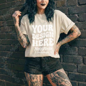 May include: A beige t-shirt with rolled-up sleeves featuring the text "YOUR DESIGN HERE" in a retro font. The shirt is part of a clothing mockup, perfect for showcasing designs. The image also includes the text "MoxxyMocks" and "Vibes That Standout".