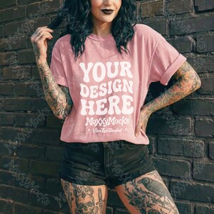 May include: A light pink t-shirt with the text "YOUR DESIGN HERE" in white, along with the brand name "MoxxyMocks" and the tagline "+ Vibes That Standout +". The shirt is paired with black shorts.