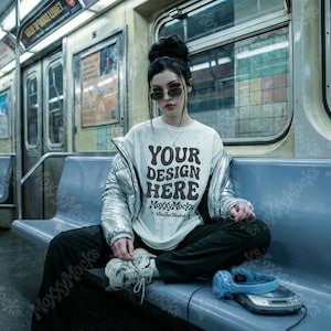 May include: A person wearing a white t-shirt with the text "YOUR DESIGN HERE" in a subway car. The person is wearing a silver jacket, black pants, and sunglasses. Headphones and a music player are on the seat.