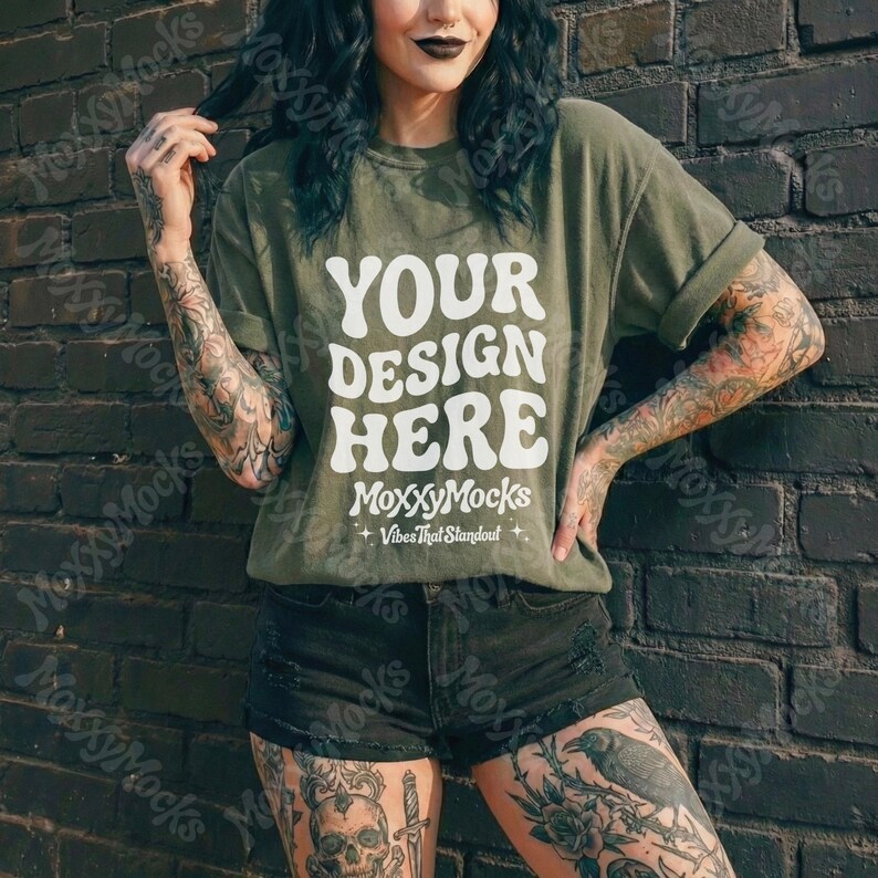 May include: Olive green t-shirt with the text "YOUR DESIGN HERE" in white, along with the brand name "MoxxyMocks" and the tagline "Vibes That Standout". The shirt is paired with black denim shorts. The model is standing in front of a dark brick wall.