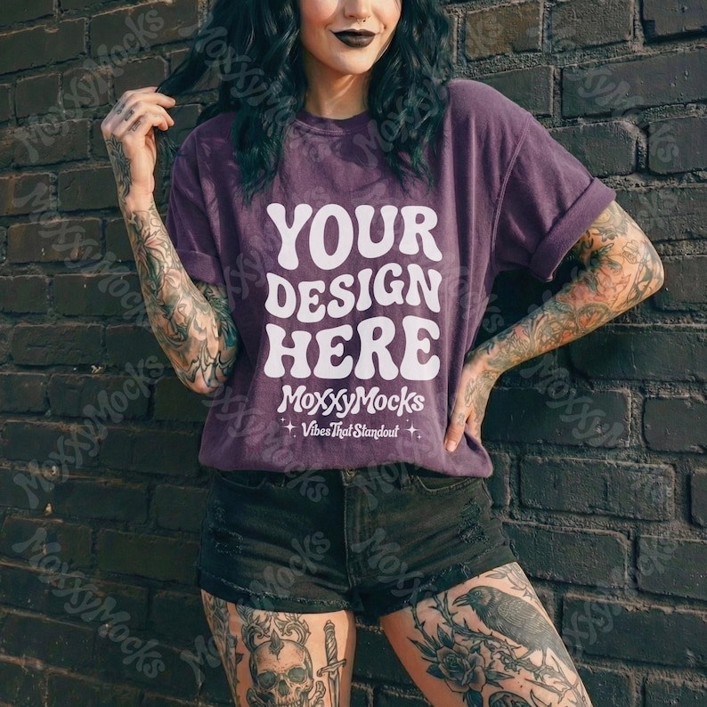 May include: A purple t-shirt with the text "YOUR DESIGN HERE" in white, with "MoxxyMocks" and "Vibes That Standout" below. The model is wearing black shorts and is leaning against a dark brick wall.