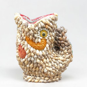 Peut inclure: Un récipient décoratif en forme de hibou, entièrement recouvert de petits coquillages clairs. Il présente un œil jaune vif, un bec brodé orange et un accent circulaire orange brodé sur le côté, avec un bord intérieur de couleur rouge.