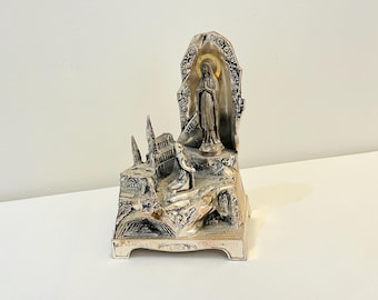 May include: A silver-toned religious statue depicting a figure with a halo, standing in front of an ornate archway. The base features architectural details and a landscape scene. The statue is on a white surface.