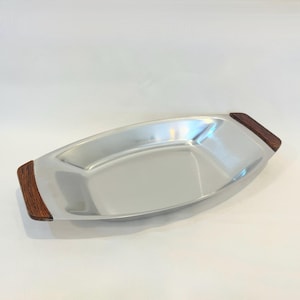 Peut inclure: Un plateau de service en acier inoxydable avec une finition polie. Le plateau a une forme ovale allongée avec des poignées en bois foncé à chaque extrémité. Le plateau est vide et repose sur une surface blanche.