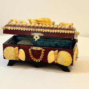 May include: A wooden box decorated with seashells. The box is open and has a blue velvet lining. The lid of the box is also decorated with seashells.