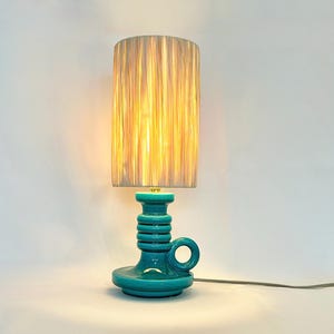 Può includere: Lampada da tavolo in ceramica turchese con paralume cilindrico. La base della lampada ha un manico e un design a coste. Il paralume è di colore beige chiaro. La lampada è collegata.