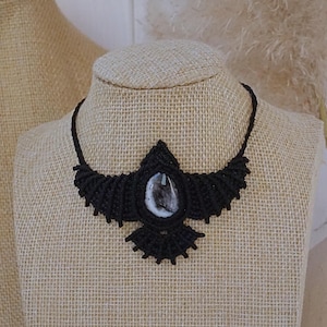 Puede incluir: Un collar de macramé negro con un colgante de piedra de ágata blanca en forma de pájaro. El collar se muestra en un maniquí cubierto de tela.