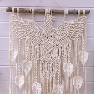 May include: A white macrame wall hanging featuring a large eagle design and smaller leaf motifs. The eagle is crafted from white cord and has an intricate pattern. The leaves are also made from white cord and hang from the bottom of the eagle.