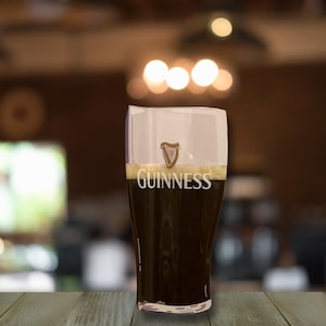Op de afbeelding: Een pintglas gevuld met donker stoutbier, bedekt met een romige, witte schuimkraag. Het Guinness-logo, een harp, en de merknaam staan op het glas. Het glas staat op een houten oppervlak, met een wazige achtergrond.
