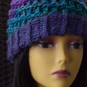 May include: A purple and teal knitted hat with a sparkly texture. The hat is worn on a mannequin with long black hair.