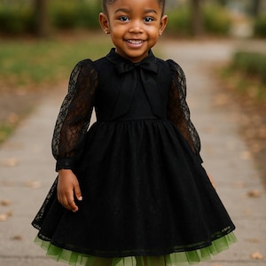Puede incluir: Una niña pequeña con un vestido de encaje negro con un dobladillo de tul verde y zapatos negros. El vestido tiene mangas largas de encaje y un lazo en el escote. Su cabello está peinado en un moño con un lazo blanco.