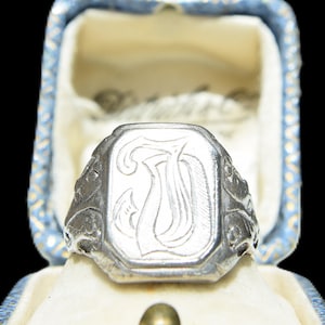 May include: A silver-toned signet ring featuring an octagonal face with an engraved letter D in a decorative script font. The ring band displays intricate embossed detailing on the sides and is presented in a blue and white patterned jewelry box.