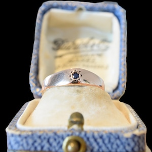 Peut inclure: Une bague en or rose avec un motif en forme d'éclat d'étoile autour d'une pierre précieuse bleu foncé, présentée dans un écrin à bijoux bleu vintage avec un intérieur crème. La bague est au centre, mettant en valeur son design élégant.