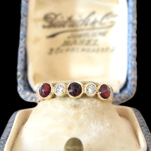 Könnte beinhalten: Ein goldener Ring mit fünf Edelsteinen: zwei rote, zwei klare und ein dunkelroter. Der Ring ist in einem goldenen Band gefasst und wird in einer Vintage-Schmuckschatulle in Blau und Creme präsentiert.