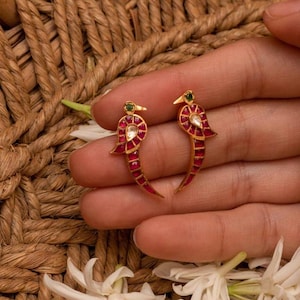 May include: A pair of gold-toned earrings shaped like birds, featuring red stone inlays along the body, a white teardrop-shaped stone at the center, and a small green stone accent at the head, designed with a curved, elongated silhouette.