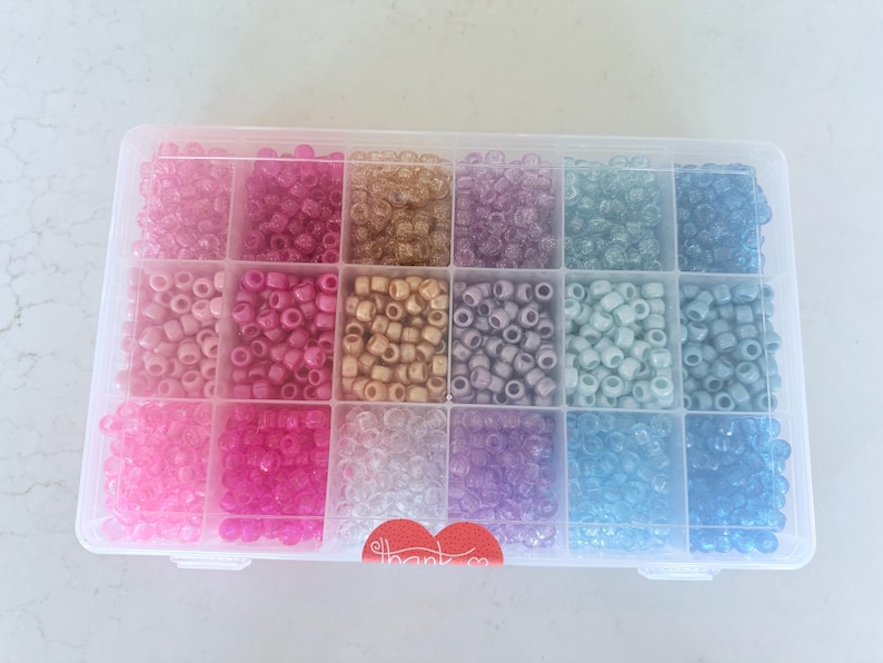 Może przedstawiać: Przezroczysty plastikowy pojemnik do przechowywania wypełniony 12 przegr&oacute;dkami z kolorowymi, tęczowymi koralikami. Koraliki są w odcieniach r&oacute;żowego, fioletowego, niebieskiego i złotego.