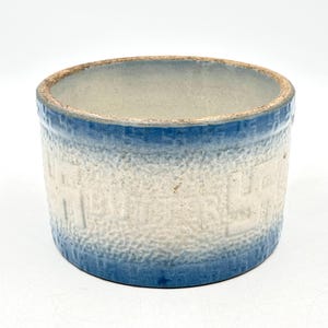 May include: A cylindrical ceramic butter crock featuring a textured, off-white body with embossed lettering that reads BUTTER. The rim and base are finished with a deep blue glaze that fades into the center, creating a distinct two-tone appearance.