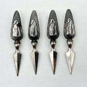 May include: Four silver-toned corn-shaped holders with a textured kernel pattern on the handle and a smooth, pointed metal prong. Each handle features a decorative embossed design of a single corn cob, providing a grip for these dining utensils.