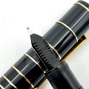 Puede incluir: Primer plano de una pluma estilogr&aacute;fica negra con detalles dorados. Se ve el plum&iacute;n del bol&iacute;grafo, con su intrincado dise&ntilde;o. Un instrumento de escritura cl&aacute;sico.