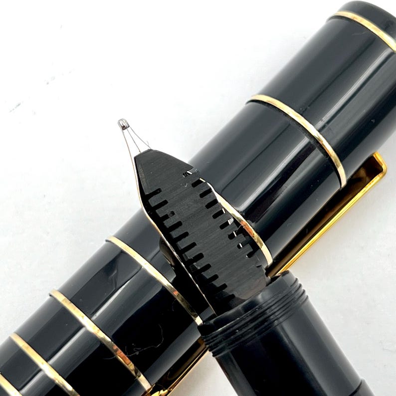 Puede incluir: Primer plano de una pluma estilogr&aacute;fica negra con detalles dorados. Se ve el plum&iacute;n del bol&iacute;grafo, con su intrincado dise&ntilde;o. Un instrumento de escritura cl&aacute;sico.