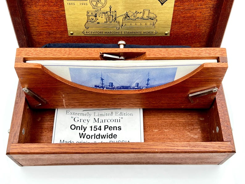 Puede incluir: Caja de madera abierta con tapa con bisagras, que revela una pluma y una imagen impresa de un barco. La caja tiene una placa de lat&oacute;n y una etiqueta que dice "Grey Marconi" y "Only 154 Pens Worldwide."