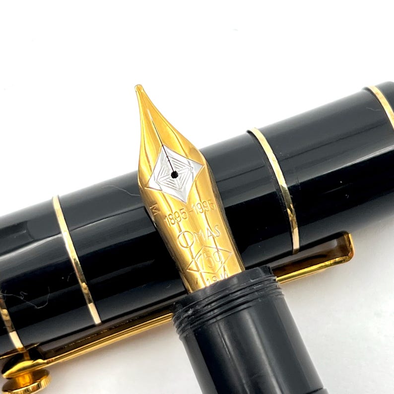 Puede incluir: Primer plano de una pluma estilogr&aacute;fica negra con detalles dorados. El plum&iacute;n dorado est&aacute; grabado con el texto "OMAS 1895-1995". La pluma tiene un cuerpo negro brillante con bandas doradas. El plum&iacute;n es dorado con un dise&ntilde;o de diamante plateado.