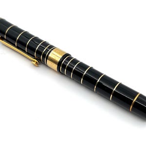 Puede incluir: Pluma estilogr&aacute;fica negra y dorada con clip dorado. El bol&iacute;grafo tiene bandas doradas alrededor del cuerpo negro. Un instrumento de escritura.