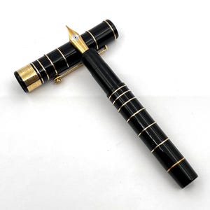 Puede incluir: Una pluma estilogr&aacute;fica negra con detalles dorados. La pluma tiene un plum&iacute;n dorado y bandas doradas alrededor del cuerpo y el capuch&oacute;n. El capuch&oacute;n est&aacute; quitado, mostrando el plum&iacute;n. La pluma est&aacute; sobre una superficie blanca.