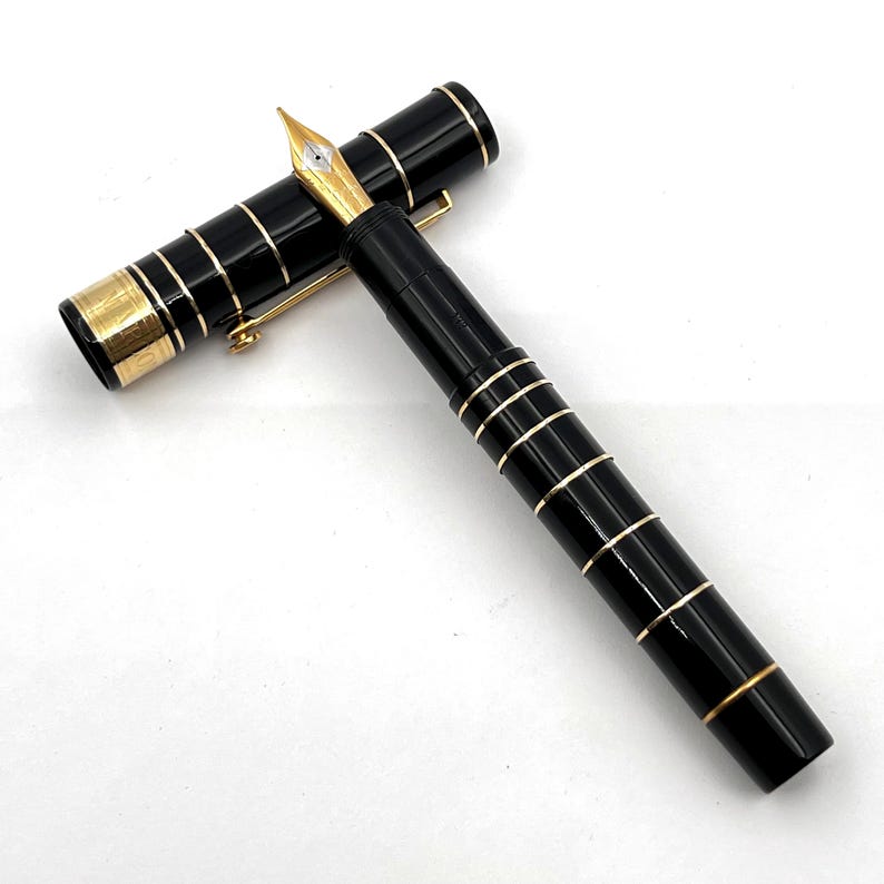 Puede incluir: Una pluma estilogr&aacute;fica negra con detalles dorados. La pluma tiene un plum&iacute;n dorado y bandas doradas alrededor del cuerpo y el capuch&oacute;n. El capuch&oacute;n est&aacute; quitado, mostrando el plum&iacute;n. La pluma est&aacute; sobre una superficie blanca.