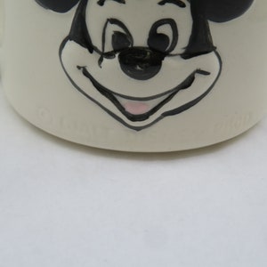 May include: White ceramic mug with a black and white illustration of Mickey Mouse's face. The mug has the word "Disney" printed on the bottom.